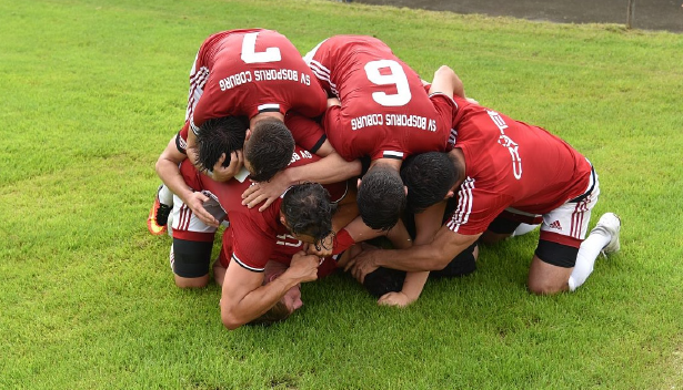 Fußballmannschaft feiert gemeinsam ein Tor, Spieler in roten Trikots liegen jubelnd übereinander