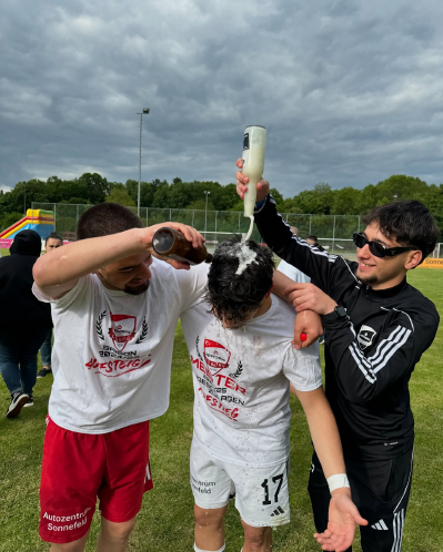 Drei Fußballspieler des SV Bosporus Coburg feiern den Aufstieg; einer bekommt Flüssigkeit über den Kopf gegossen.