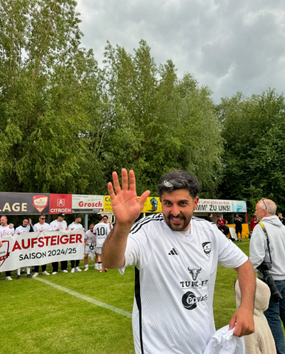 Fußballspieler des SV Bosporus Coburg winkt jubelnd in die Kamera, im Hintergrund steht das Team mit einem Aufstiegsbanner.